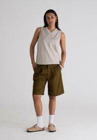 Beige, ujj nélküli, V-nyakú felső, olívazöld, redőzött shorts, fehér zoknik és világosbarna slip-on cipők, egyszerű háttér előtt.
