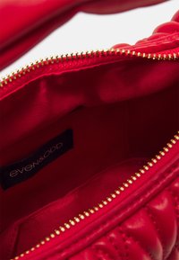 Sac à main rouge matelassé ouvert montrant l'intérieur en tissu rouge lisse et une petite étiquette noire avec le nom de la marque even&odd à l'intérieur.