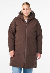 Femme portant un manteau d'hiver à capuche marron avec une fermeture éclair à l'avant et un jean bleu clair, se tenant devant un fond blanc uni.
