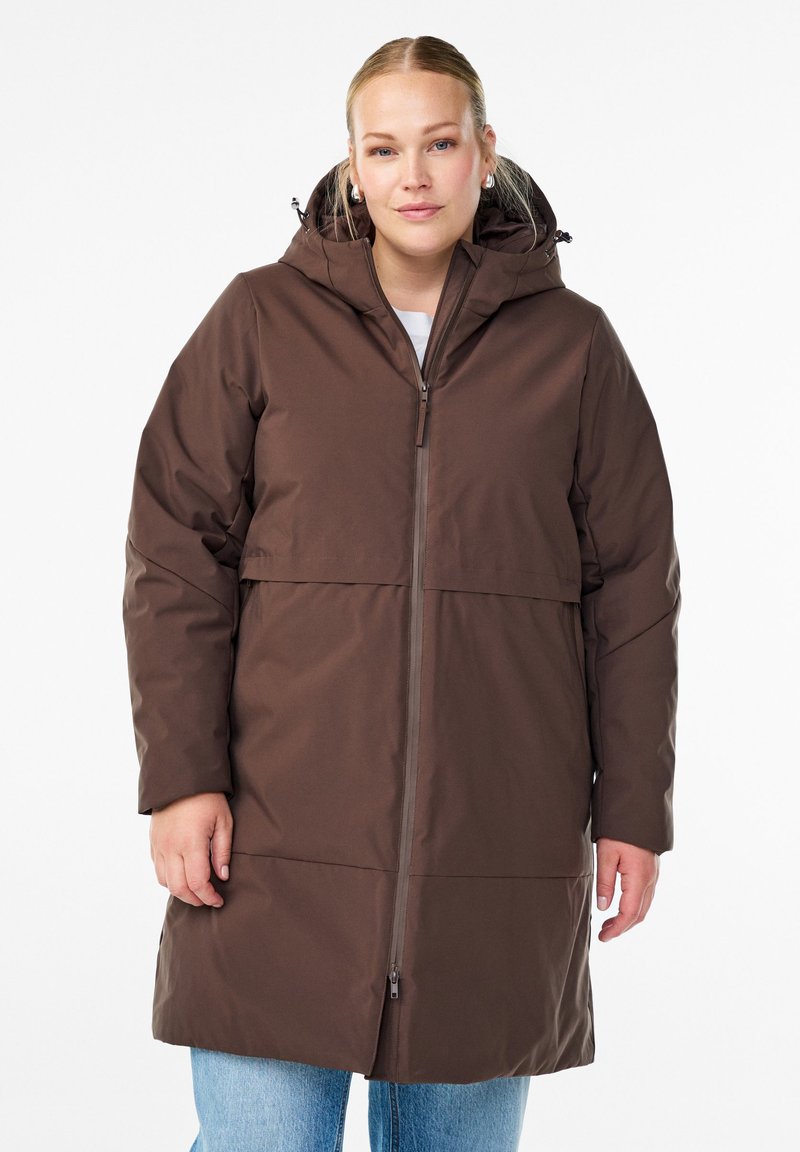 Femme portant un manteau d'hiver à capuche marron avec une fermeture éclair à l'avant et un jean bleu clair, se tenant devant un fond blanc uni.