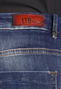 Jean en denim bleu foncé avec une surface texturée, arborant une étiquette en cuir marron LTB, des coutures jaunes et des détails sur la poche arrière.