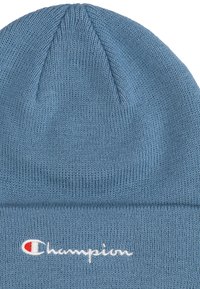 Gorro de punto azul con diseño acanalado. Presenta el logo bordado de "Champion" en blanco y rojo en el borde doblado. Textura suave.