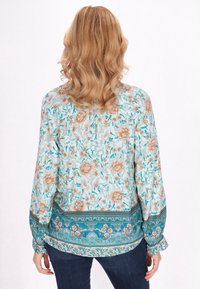 Bluză florală de culoare albastră deschisă, cu modele de flori piersici și verzi. Mâneci lungi, manșete gathered și o margine cu model.