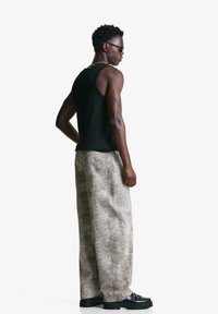 Schwarzes Tanktop, beiges strukturiertes weites Jeansmodell und schwarze Schnürschuhe. Mann steht mit dem Rücken zur Kamera, um die Details und Passform des Outfits zu zeigen.