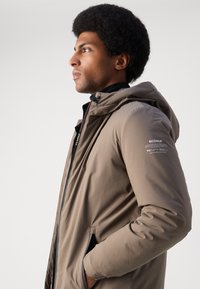 Chaqueta con capucha marrón y textura suave. Presenta bolsillos laterales, capucha ajustable con cordón y la marca Ecoalf en la manga.