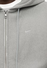 Γκρι φόρμα με φερμουάρ κατασκευασμένη από μαλακό ύφασμα. Διαθέτει ασημένιο φερμουάρ, κορδόνι και μικρό λογότυπο Nike σε λευκό στο στήθος.