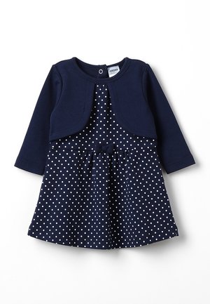 Vestido azul marino con lunares blancos, mangas largas y una chaqueta corta a juego. Presenta un lazo en la cintura y un escote redondo.