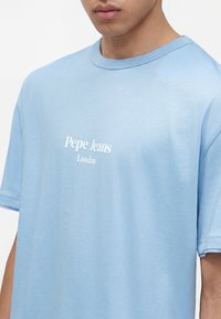Άνδρας που φοράει ένα ανοιχτό μπλε T-shirt Pepe Jeans London με κοντά μανίκια και στρογγυλή λαιμόκοψη, εμφανιζόμενος από το στήθος έως τον λαιμό.