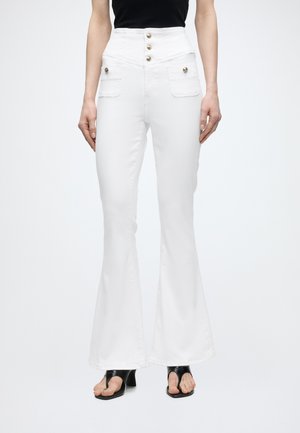 Γυναίκα που φοράει λευκό παντελόνι με ψηλή μέση, καμπάνα, με μπροστινές τσέπες και χρυσά κουμπιά, συνδυασμένο με μαύρα πέδιλα με τακούνι και ανοιχτά δάχτυλα και μαύρο τοπ.