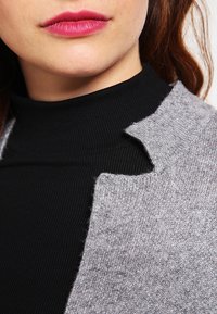 Grijze gebreide cardigan met een geribde zwarte coltrui eronder. Opvallende kraagvorm met een lichte uitsnijding bij de halslijn.