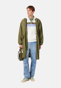 Chaqueta larga verde oliva sobre una sudadera con capucha blanca con rayas azules y verdes, combinada con vaqueros azul claro y zapatillas verdes, sosteniendo una bolsa gris.