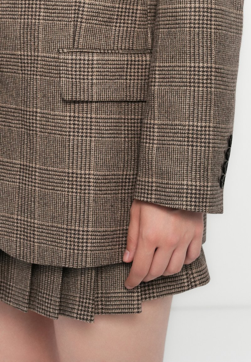 Blazer a quadri marrone con un design strutturato, dotato di una singola tasca sul petto. Gonna plissettata a strati con motivo a quadri coordinato.