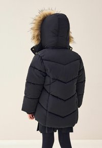Doudoune noire avec une capuche en fausse fourrure. Design en chevrons, tissu épais et accents dorés sur les boutons. Vue arrière montrée.