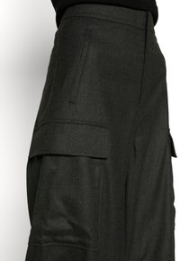 Pantalon cargo gris foncé taille haute avec poches à rabat latérales et une poche verticale fendue, porté avec un haut noir.