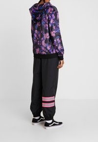 Person som bär svarta vida byxor med rosa reflexremmar och en lila blommönstrad huvtröja, stående mot en enfärgad vit bakgrund.