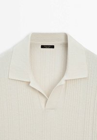 Pull en maille crème avec un motif côtelé, un col doux et un col en V ouvert, montrant une étiquette en tissu tissé à l'intérieur de l'encolure.