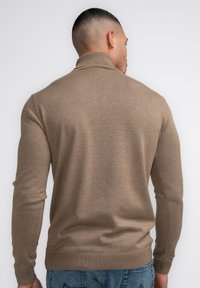 Brauner Pullover mit hohem Kragen, Rippbündchen und glatter Textur. Getragen mit blauen Jeans, von hinten betrachtet.