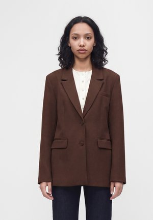 Mujer con cabello rizado y oscuro lleva una blazer marrón sobre una camisa blanca abotonada y pantalones oscuros, de pie frente a un fondo blanco liso.