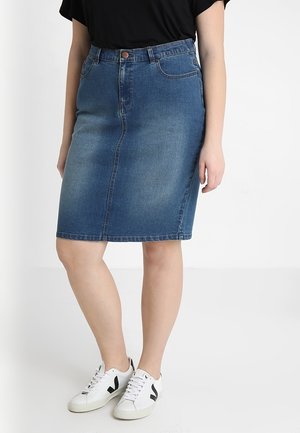 Frau trägt einen knieumspielenden blauen Jeansrock, schwarzen Oberteil und weiße Sneaker mit schwarzem Logo, steht vor weißem Hintergrund.