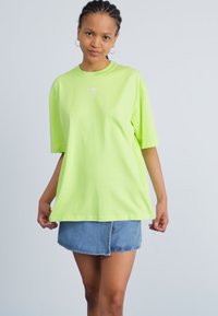 Neónovo zelené oversized tričko s krátkymi rukávmi, s malým bielym logom na hrudi. V kombinácii s modrou džínsovou mini sukňou.