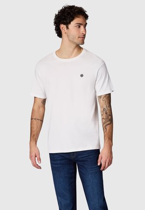 Mężczyzna z kręconymi włosami, ubrany w białą koszulkę z małym czarnym logo koła sterowego statku, niebieskie dżinsy i tatuaże na obu przedramionach, patrzący w prawo.