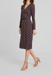 Långärmad klänning med en djup V-halsringning, omslag design och ett färgglatt geometriskt mönster på en mörk bakgrund. Bärs med tan blockklackar.