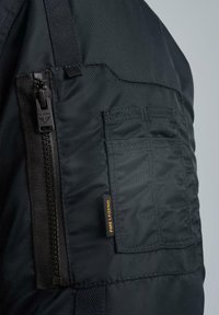 Schwarze gepolsterte Jacke mit einem seitlichen Reißverschluss, mehreren Taschen und einem Etikett mit der Aufschrift "PME LEGEND". Der Stoff wirkt glatt und langlebig.