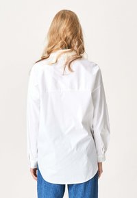 Person med langt blondt hår iført en løs hvid skjorte med knapper og blå jeans, vist set fra ryggen foran en ensfarvet baggrund.