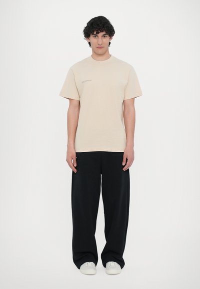 Beige cotton t-shirt with short sleeves and a crew neckline, featuring minimal text detail. Paired with loose-fitting black trousers and white sneakers.
