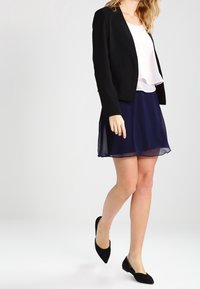 Černý blazer přes bílou blůzu, spárovaný s námořnickou chiffonovou sukní a černými balerínami, ukazuje vrstvený design a hladkou texturu.