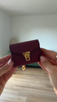 Μικρό χαλινού wallet από μπορντό δέρμα με κλείσιμο τύπου καπάκι, διαθέτοντας χρυσό μεταλλικό κούμπωμα και προσάρτηση για κλειδιά. Υφή επιφάνειας, συμπαγής σχεδίαση.