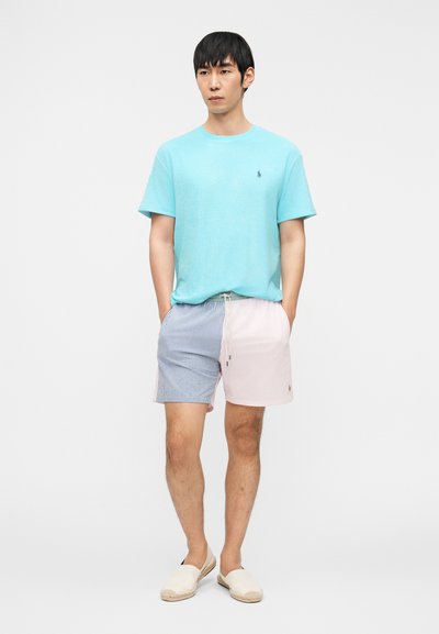 Άνδρας που φοράει πουκάμισο με κοντά μανίκια σε τιρκουάζ χρώμα, σορτς δίχρωμα σε ανοικτό μπλε και ροζ, και μπεζ παπούτσια slip-on, όρθιος με τα χέρια στις τσέπες.