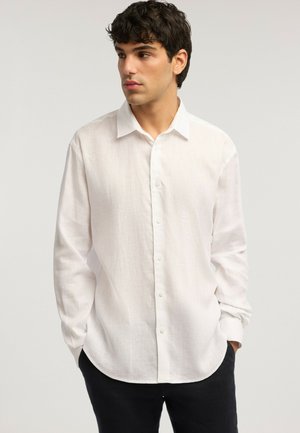 Junger Mann mit dunklen Haaren, der ein langärmliges weißes Hemd mit Knöpfen und schwarze Hose trägt, schaut nach links und hat die Hände in den Taschen.