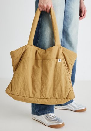 Hånd, der holder stor beige taske, båret med blå jeans og hvide sneakers på en ensfarvet baggrund.