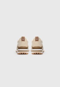 Sneaker beige e bianche con tiranti marroni numerati "1967," viste da dietro, mostrando suole in gomma dotate di battistrada su uno sfondo bianco.
