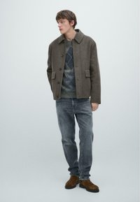 Cappotto di lana marrone con grandi tasche e colletto, indossato sopra un maglione argyle grigio e blu, abbinato a jeans grigio chiaro e stivali marroni.