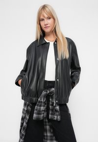 Veste bomber en simili cuir noir avec un col, fermeture zippée à l'avant, poignets élastiques et ourlet ; portée sur un haut blanc et une chemise à carreaux.