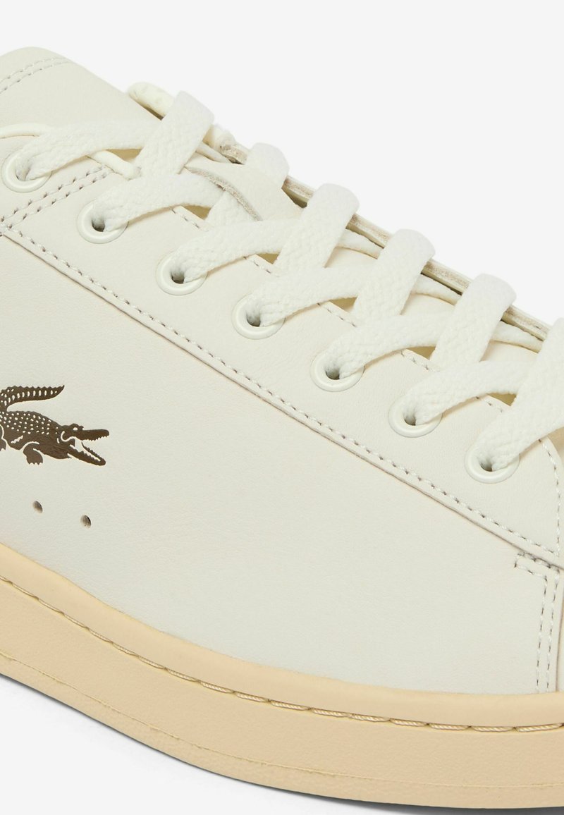 Primo piano di una sneaker in pelle bianca con lacci bianchi, suola beige e un piccolo logo scuro a forma di coccodrillo sul lato.