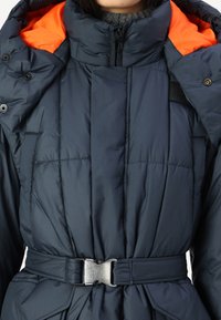 Giacca imbottita blu navy con colletto interno arancione, vita con cintura e dettagli a bottoni a pressione; presenta una texture trapuntata e zip regolabile.