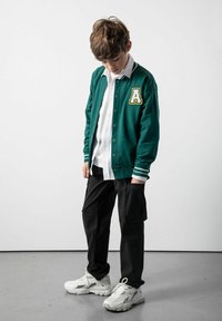 Zielona kurtka typu varsity z białymi paskami, wyszywanym 'A', noszona na białej koszuli i czarnych spodniach cargo, w połączeniu z białymi trampkami.
