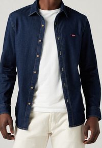 Camicia in denim blu scuro con chiusura a bottoni, caratterizzata da accessori in oro e una toppa con logo rosso. Indossata sopra una T-shirt bianca.