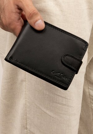 Hand holding a closed black leather wallet with snap button over beige fabric clothing background.