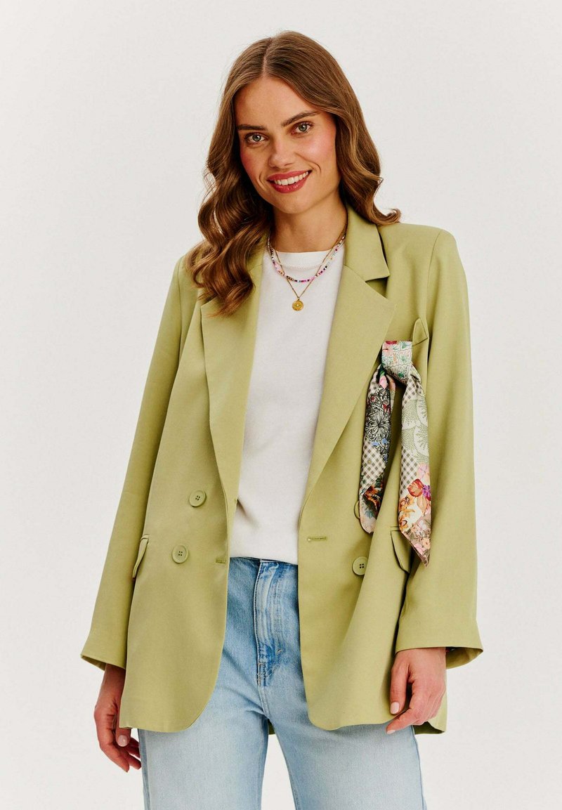 Jasnozielona marynarka dwurzędowa o nieco oversize'owym kroju, z dwoma guzikami i kwiatowym akcentem w kieszeni.