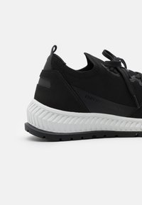 Svarta sportskor med en texturerad stickad ovandel, mockadetaljer, tjock gummisula och en vit panel med zigzagmönster. Logotypen för varumärket på sidan.