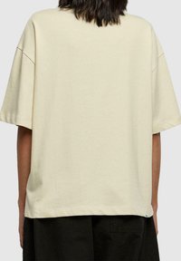 Camiseta beige de gran tamaño en algodón suave, con mangas cortas y cuello redondo. El dobladillo es recto con detalles de costura mínimos.