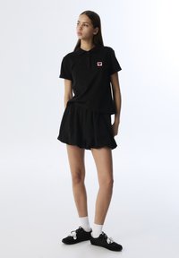 Camiseta polo negra con un parche en forma de corazón, combinada con shorts negros. Tejido suave, corte relajado, mangas cortas, dobladillo redondeado, llevada con zapatillas negras.