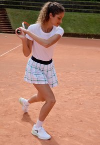 Maglietta sportiva bianca abbinata a una gonna a quadretti bianca, verde e blu. Sneakers bianche con dettagli verdi. Su un campo da tennis in terra rossa.