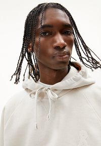 Felpa con cappuccio di colore beige chiaro, realizzata in tessuto morbido. Presenta un cappuccio con cordoncino, dettagli a coste e un logo ricamato sul davanti.