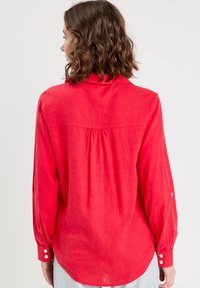 Rote Bluse mit langen Ärmeln aus leichtem Stoff, mit einem Faltenwurf auf dem Rücken, abgerundetem Saum und Knopfakzenten an den Bündchen.