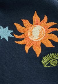 Tessuto ricamato con un sole arancione brillante, una stella azzurra chiara e un disegno di occhio verde su uno sfondo blu scuro. Tessuto testurizzato.